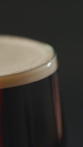 Vertical-Video-Close-Up-Of-Pint-Of-Irish-Stout-In-Glass-Against-Black-Background-To-Celebrate-St-Patricks-Day